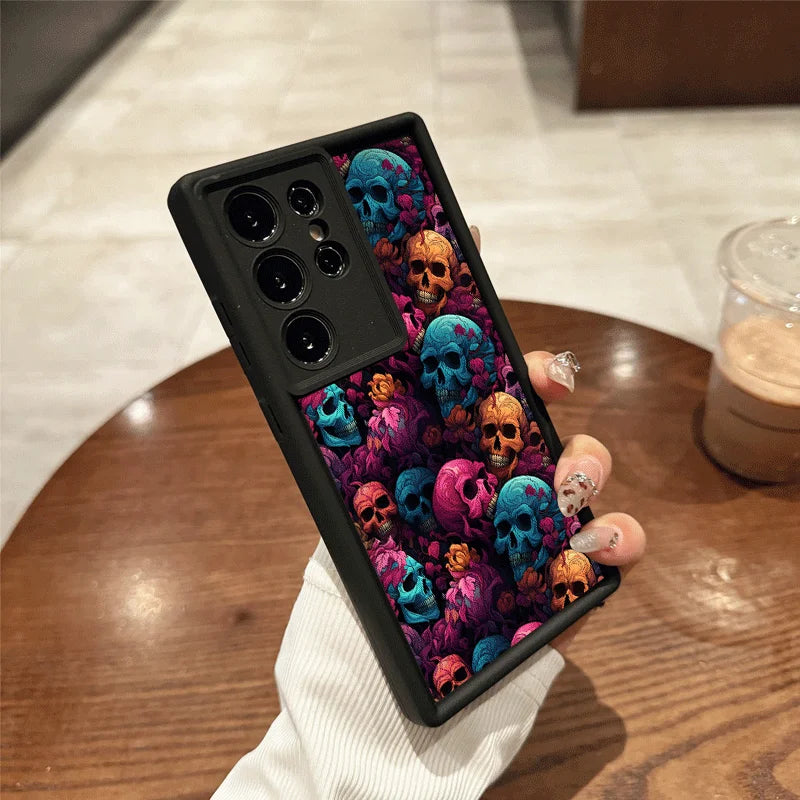 Hand hält schwarze Samsung-Hülle mit mehrfarbigen Totenköpfen in Blau, Orange und Pink auf dunklem Blumenhintergrund